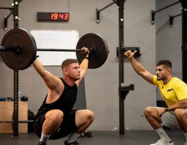Athlete performing weightlifting exercise under trainer's guidance in a gym setting.